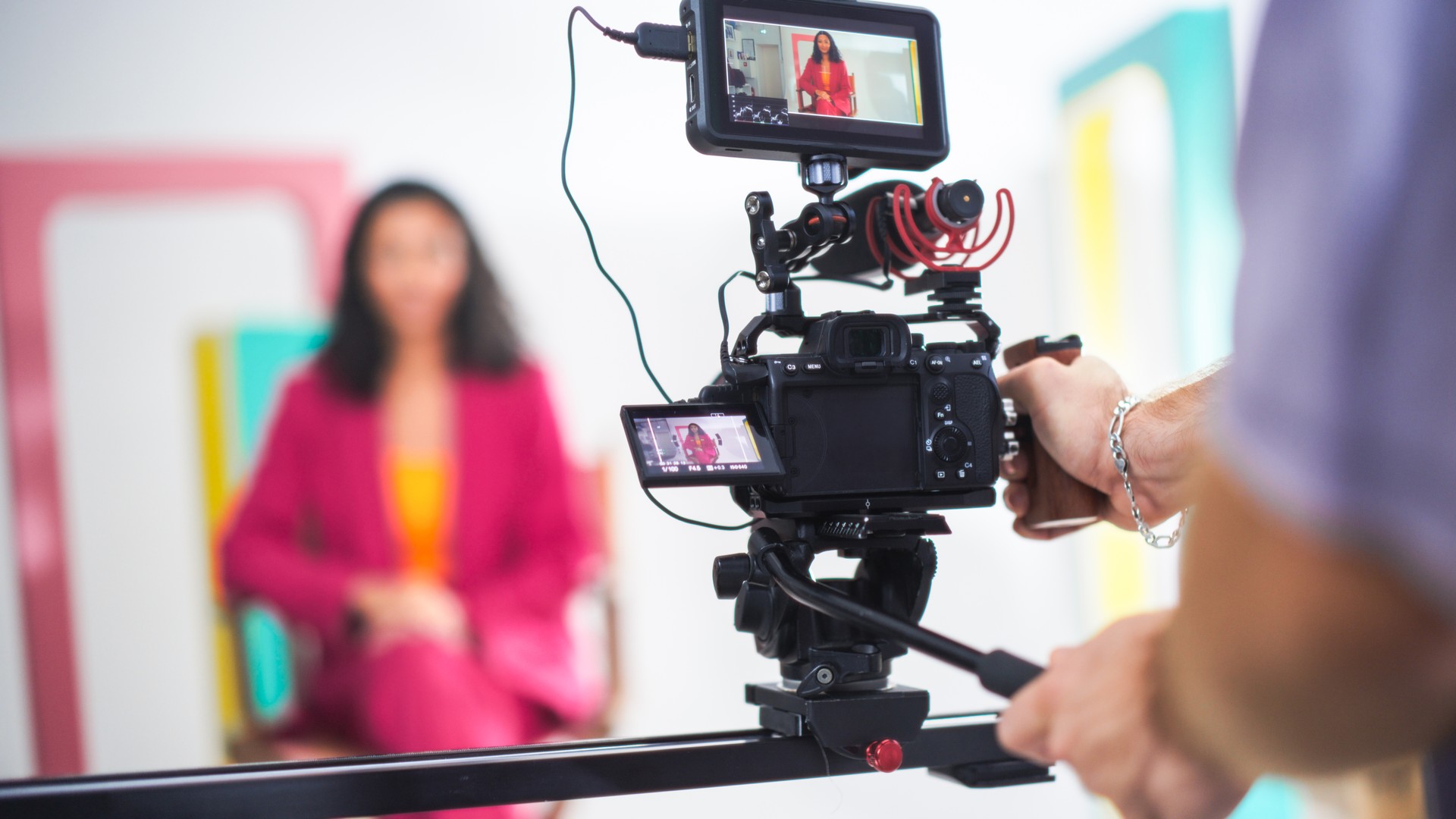 Una escena vibrante captura a una joven anfitriona asiática con un blazer rosa en el set de filmación, participando en una animada discusión, con un enfoque en el equipo de video profesional y el diseño dinámico del escenario.